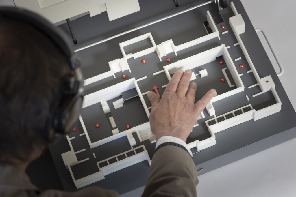 Het model is open geklapt. Een man verkent met zijn rechterhand de aula van het museum en luistert naar de audio via de koptelefoon