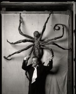 Fotobeschrijving: portret van Louise Bourgeois met boven haar hoofd een van haar kunstwerken, een spin. 