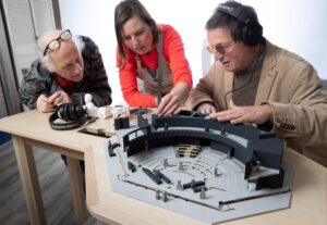 Blinde man luistert met koptelefoon naar de audiomaquette terwijl twee andere personen de maquette inspecteren.